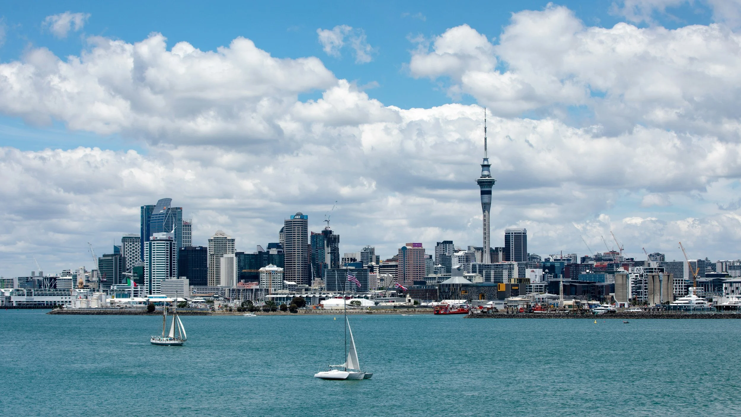 City skyline with modern skyscrapers, including the Sky Tower, seen across a body of water with sailboats and a partly cloudy sky.